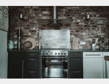 Moderne Küche mit Ziegelsteinwand im Hintergrund, zentralem Edelstahlherd mit Dunstabzugshaube und schwarzen sowie weißen Unterschränken. Auf der Arbeitsfläche befinden sich Küchenutensilien wie Schneidebretter, ein Wasserkocher und eine Vase mit Dekoration.