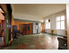 Ein einfacher Innenraum mit einer abgenutzten Bodenfläche, Holzwandverkleidung und hohen weißen Wänden. Der Raum enthält Schließfächer, einen schwarzen Klavierstuhl, antike Schränke mit farbigen Scheiben, eine Leiter und große Fenster, die natürliches Licht hereinlassen.