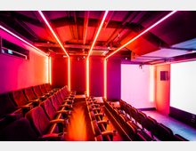 Kinosaal mit Reihen gepolsterter Kinosessel, rotem und pinkem Neonlicht an den Wänden und der Decke sowie einer weißen Leinwand an der Vorderseite. Die Decke ist mit sichtbaren Rohren und technischen Elementen gestaltet.