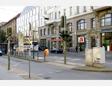 Straßenszene in einer Stadt mit Mehrfamilienhäusern im Hintergrund, darunter ein modernes Gebäude mit einer Glasfassade und ein historisches Gebäude mit Details aus Stein. Im Vordergrund befindet sich eine Bushaltestelle, Straßenlaternen und mehrere geparkte Fahrzeuge, darunter ein Reisebus und ein Auto.