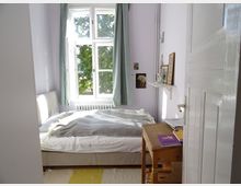 Kleines Schlafzimmer mit einem Einzelbett, das vor einem großen Fenster mit grünen Vorhängen steht. Rechts befindet sich ein kleiner Holztisch mit Büchern, dekorativen Gegenständen und gerahmten Bildern an der Wand.
