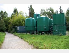 Industrieanlage mit mehreren großen, grünen Tanks, umgeben von einem Zaun, in einer grünen, bewaldeten Umgebung. Im Vordergrund führt ein gepflasterter Weg entlang einer Wiese.