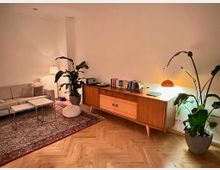 Gemütlicher Wohnbereich mit hellgrauem Sofa und zwei Couchtischen auf einem gemusterten Teppich sowie Parkett im Fischgrätmuster. Daneben steht ein niedriges Holz-Sideboard mit Büchern und Lampe, flankiert von großen Zimmerpflanzen vor einer schlichten weißen Wand.