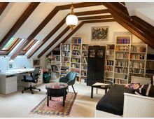 Ein Arbeitszimmer unter einer Dachschräge mit freiliegenden Holzbalken, ausgestattet mit einem Schreibtisch, einem schwarzen Bürostuhl und Dachfenstern auf der linken Seite. Im Raum befinden sich Bücherregale voller Bücher, ein Tagesbett, ein runder Beistelltisch mit Teppich sowie eine Hängelampe in der Mitte.