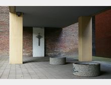 Überdachter Innenhof mit Pflastersteinen, gelben Säulen und freien runden Betonpflanzkübeln. An einer weißen Wand zwischen zwei Klinkerwänden hängt ein schmiedeeisernes Kreuz.