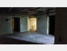 Ein leerer, dunkler Kellerraum mit weißen, teilweise beschädigten Wänden aus Ziegeln. Im Hintergrund sind geöffnete Türen zu erkennen, durch die schwaches Licht einfällt; freiliegende Rohre und Kabel verlaufen unter der Decke.