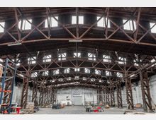 Weitläufige Industriehalle mit Holzträgerkonstruktion, einer hohen Decke und großen Oberlichtern, die natürliches Licht einlassen. Der Raum enthält diverse Lagergegenstände, Paletten und Regale, die entlang der weißen Wände verteilt sind.