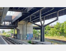 Bahnhofsumgebung mit Gleisen, überdachter Plattform und Brückenkonstruktion aus Stahlträgern. Im Hintergrund sind Betonpfeiler, Graffiti und angrenzende Grünflächen sichtbar.