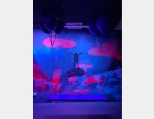 Ein surreal gestalteter Innenraum mit blau-violetter Beleuchtung, einer Wandmalerei aus leuchtenden Pilzen und einer Silhouette einer Person auf einem Felsen. Im Vordergrund schwebt eine Schaukel an Ketten, dekoriert mit schwarzen Ballons.