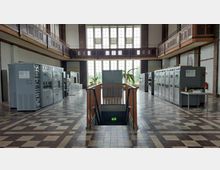 Historische technische Anlage mit großen, grauen Schaltschränken in einem hohen Raum mit Fensterfront, umlaufender Holzbalkon-Galerie und gemustertem Fliesenboden. In der Mitte des Raumes befindet sich ein Treppenabgang mit Holzgittergeländer und Notausgangszeichen.