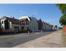 Straßenszene mit einer Reihe von Wohnhäusern, darunter modernisierte und historische Gebäude mit Satteldächern, entlang einer asphaltierten Straße mit Baustellenmarkierungen. Rechts verläuft ein Gehweg mit Kopfsteinpflaster, daneben eine rote Backsteinmauer und Bäume.