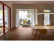 Ein heller Innenraum mit Holzboden, großen Fenstern mit weißen Vorhängen und einer Glastür mit rotem Rahmen. Im Vordergrund steht ein langer weißer Tisch mit gelben Stühlen, und durch die Fenster sind eine Straße, parkende Autos und ein gelbes Lastenrad sichtbar.