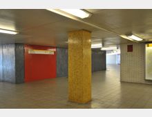 Unterirdischer Durchgang mit gefliesten Wänden in verschiedenen Farben: ein gelber Mosaikpfeiler, eine rote Wand mit Wegweisern und weiße sowie blaue Fliesen. Der Bereich ist mit Deckenleuchten beleuchtet.