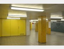 Unterirdischer Durchgang mit gelben Wandpaneelen, gelben Fliesenstützen und weißen Ziegelwänden. Die Decke ist mit langen Leuchtstofflampen beleuchtet, der Boden besteht aus grauen Fliesen.