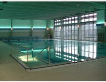 Hallenbad mit einem rechteckigen Schwimmbecken, umgeben von glatten Fliesen. Große Fenster an einer Wand lassen Tageslicht herein, und die Innenbeleuchtung in Türkistönen unterstreicht die moderne Einrichtung.