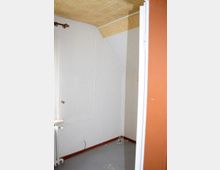 Ein kleiner, leerer Raum mit weißer Tapete, einer schrägen Decke mit Holzverkleidung und grauem Boden. An der Wand sind sichtbare Kabel und eine Steckdose, und ein Heizkörper sowie ein Fenster mit einer weißen Gardine befinden sich links.