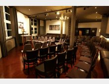 Innenansicht eines kleinen Veranstaltungs- oder Vortragsraums mit vielen in Reihen aufgestellten Stühlen, einer kleinen Bühne mit Rednerpult und großen Fenstern mit Vorhängen an der linken Wand. Der Raum hat eine hohe Decke mit Spotbeleuchtung und Kronleuchter, Säulen, dunkle Sitzbänke an den Wänden und einen rötlichen Boden.