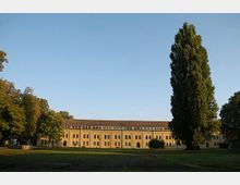 Historisches Backsteingebäude mit zahlreichen Fenstern und einem flachen Dach vor einer großen, offenen Wiese; rechts ein hoher, schmaler Baum und links sowie rechts weitere Bäume am Rand des Geländes.