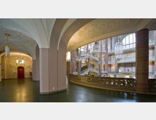 Innenansicht eines historischen Gebäudes mit geschwungenen Treppen, verzierten Geländern und hohen Rundsäulen. Die Wände und Decken sind mit floralen Stuckmustern verziert, große Fenster lassen Tageslicht in die Halle, und im Vordergrund ist eine rote Tür mit dekorativen Ornamenten sichtbar.