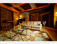 Gemütlicher Raum mit Holzwänden und einem Steinakzentbereich. Im Vordergrund steht ein gläserner Couchtisch auf einem gemusterten Teppich, dahinter befindet sich ein beiges Sofa und ein Klavier, das in der Ecke neben einer Tür und einer Lampe steht.