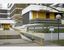 Moderner Gebäudeeingang mit gelben Türen und gelben Handläufen an Treppen und Rampen. Die Fassade besteht aus einer Kombination aus weißen und dunkelbraunen Elementen, mit blühenden Bäumen und einem Hinweisschild im Vordergrund.