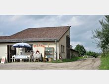 Ein schlichtes, ländliches Gebäude mit heller Fassade und Ziegeldach, vor dem ein kleiner Keramikverkauf stattfindet. Ein Tisch mit einem Sonnenschirm sowie ein Regal mit Keramikwaren stehen vor dem Eingang, daneben führt ein unbefestigter Weg entlang einer grünen Landschaft.