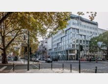 Kreuzung einer Stadtstraße mit einem modernen, mehrstöckigen Gebäude mit Glasfassade an einer Ecke; im Vordergrund Herbstbäume, Ampeln und Fußgänger. Auf der Straße stehen Autos geparkt, und Menschen sind zu Fuß unterwegs.