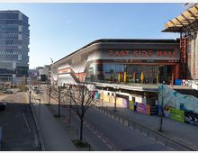 Straßenansicht eines modernen Einkaufszentrums namens "East Side Mall" mit geschwungenem, schwarz-weiß-rotem Fassadendesign. Im Hintergrund ein hohes Bürogebäude und Baukräne, im Vordergrund eine Straße mit vereinzelten Bäumen und Fahrradständern.