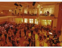 Tanzveranstaltung in einem zweistöckigen Saal mit warmem, gelbem Licht; der untere Bereich ist gefüllt mit tanzenden Paaren, während auf der Empore Menschen sitzen und zusehen. Die Wände sind mit großen Fenstern und Vorhängen gestaltet, und an der Decke hängen Beleuchtung sowie eine Diskokugel.