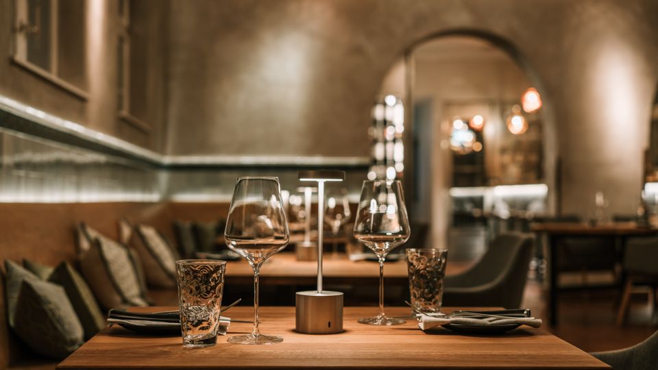 Innenraum eines modern eingerichteten Restaurants mit warmem, gedämpftem Licht, Holztischen und gepolsterten Sitzbänken an der Wand. Im Vordergrund ein gedeckter Tisch mit Weingläsern, Wassergläsern und kleiner Tischlampe; im Hintergrund ein Rundbogen-Durchgang zu weiteren Sitzbereichen.