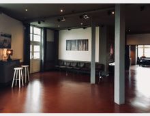 Moderner Veranstaltungsraum mit roten Böden, grauen Wänden und minimalistischer Einrichtung. Sitzbänke befinden sich entlang einer Wand, neben einer Tür mit Glasfenstern, und eine kleine Theke mit Barhockern steht links im Bild.