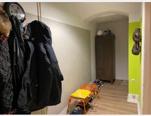 Schmaler Flur mit hellbeigen Wänden, teils grün akzentuiert. Links hängt eine Garderobe mit Jacken, daneben steht eine orange Sitzbank. Im Hintergrund befindet sich ein brauner Metallschrank, und an der Wand hängen Boxhandschuhe. Der Boden ist mit Holzoptik-Laminat ausgelegt.