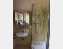Kleines Badezimmer mit beigefliesten Wänden, Waschbecken mit Spiegel- und Glasablage voller Pflegeprodukte sowie einer Eckdusche mit gebogener Glasabtrennung. Ein Fenster sorgt für Tageslicht und spiegelt sich im Spiegelschrank.