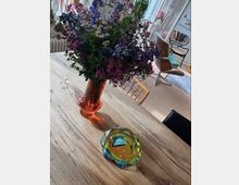 Ein Holztisch mit einer großen Vase voller bunter Blumen im Vordergrund. Dahinter sind moderne Möbel wie ein Sessel und ein heller Raum mit großen Fenstern zu sehen.