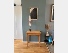 Ein kleiner Holztisch steht vor einer dunkelgrünen Wand, darauf steht eine braune Vase mit getrockneten Gräsern. An der Wand hängt ein gerahmtes Kunstwerk mit abstraktem Motiv, daneben sind weitere Dekorationselemente wie Vasen und Pampasgras zu sehen.