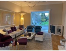 Wohnzimmer mit modernem Design, bestehend aus einem cremefarbenen Sofa, violetten und blauen Sesseln, Glastischen und einem großen Kunstwerk an der Wand. Eine große Glastür im Hintergrund öffnet sich zu einem Garten mit grünem Rasen und Bäumen.