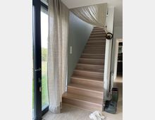 Treppenaufgang in einem modernen Wohnraum mit Holzstufen, links eine Glastür mit beigefarbenem Vorhang. Neben der Treppe stehen Gummistiefel auf einem hellen Teppich, und über der Treppe hängt ein gefalteter, transparenter Stoff als Dekoration.