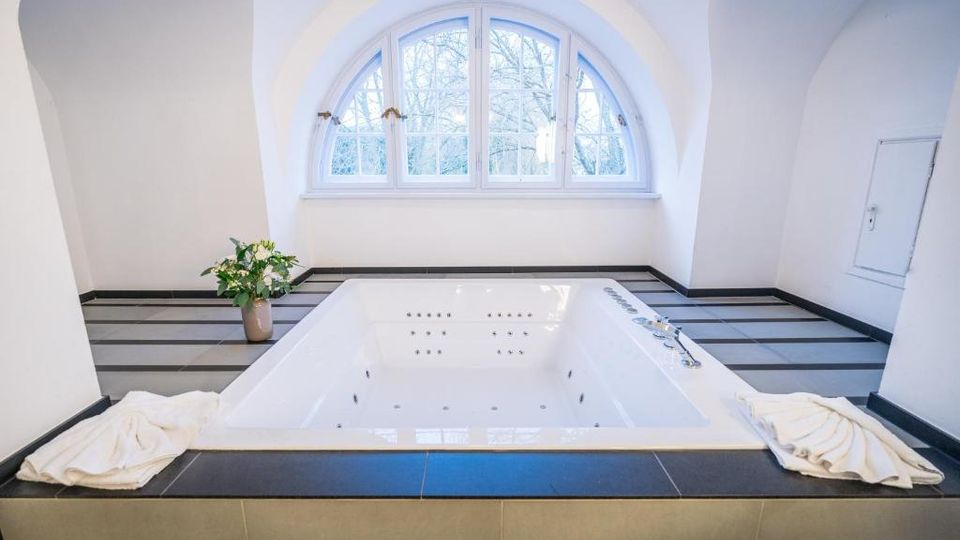 Helles Wellness-Badezimmer mit einem eingelassenen Whirlpool in gefliester Umrandung und integrierten Düsen. Dahinter befindet sich ein großes Rundbogenfenster, umgeben von weiß verputzten, gewölbten Wänden.