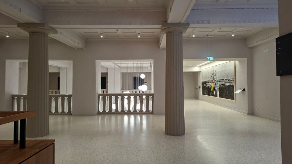Heller Museums- oder Galerieinnenraum mit hoher Kassettendecke, breiten Unterzügen und zwei großen, geriffelten Säulen. Offene Balustraden zum angrenzenden Bereich, moderne Hängelampen und ein großformatiges Wandbild an der rechten Wand.