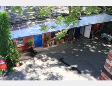 Innenhof mit einem niedrigen, langgezogenen Backsteingebäude, blau gestrichenen Türen und Fenstern sowie einem flachen, teils transparenten Dach. Vor dem Gebäude stehen Topfpflanzen; Bäume werfen Schatten auf den Asphalt, daneben sind ein Fahrrad und ein kleiner Sitzbereich zu sehen.