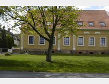 Gelbes Gebäude mit Satteldach und weißen Fensterrahmen, umgeben von einer gepflegten Rasenfläche. Ein großer Baum steht im Vordergrund, und vor dem Gebäude sind gemauerte Elemente mit Natursteinen sichtbar.