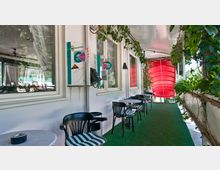 Überdachte Terrasse mit Rasenboden und Tischen mit schwarz-weiß gestreiften Stühlen, an der weißen Hauswand ein Münztelefon und Fenster; grüne Pflanzenranken und ein rotes, rundes Element im Hintergrund.