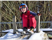 Ein Kind in rotem Schneeanzug und blauer Skibrille sitzt auf Skiern an einem schneebedeckten Holzpodest vor einem Hintergrund aus kahlen Bäumen.