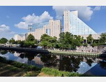 Großes modernes Hotel mit Glas- und Sandsteinfassade am Ufer eines Flusses, umgeben von Bäumen und einer Promenade mit Sitzgelegenheiten. Das Gebäude spiegelt sich im ruhigen Wasser, während der Himmel klar und leicht bewölkt ist.