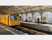 Gelber U-Bahn-Waggon mit der Aufschrift "Uhlandstraße" steht in einem überdachten Bahnhof mit großen, bogenförmigen Fenstern; auf dem gegenüberliegenden Gleisbereich sitzt eine Person auf einer Bank, weitere Personen und ein Fahrkartenautomat sind sichtbar. Im Hintergrund steht das Stationsschild „Görlitzer Bahnhof“.