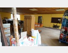 Ein Innenraum mit gelber Wandfarbe und hölzernem Kassettendach, gefüllt mit diversen Gegenständen wie Möbeln, Kartons, Säcken, einem Holzschrank und einem orangen Einkaufswagen. Der Raum wirkt unaufgeräumt, mit gelagerten Baustoffen und Haushaltsgegenständen.