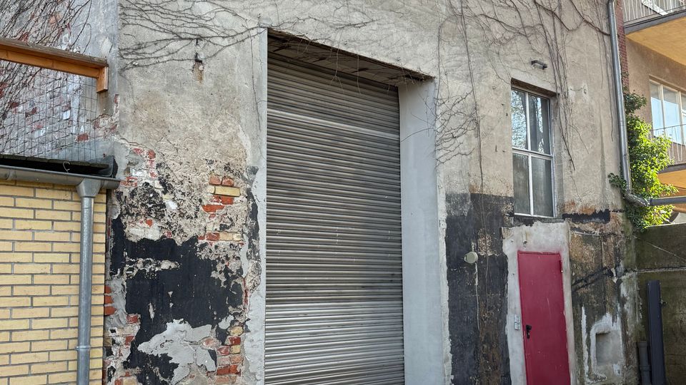 Außenansicht eines älteren Nebengebäudes mit verwitterter, teils freiliegender Ziegel- und Putzfassade, überwachsen von kahlen Kletterranken. In der Front befinden sich ein großes graues Rolltor, ein Fenster und eine kleine rote Tür; davor liegt ein gepflasterter Hof zwischen angrenzenden Wohngebäuden.