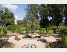 Ein gepflegter Park mit einem zentralen Springbrunnen aus dunklem Stein, umgeben von symmetrisch bepflanzten Blumenbeeten in leuchtenden Farben wie Rot, Lila und Gelb. Im Hintergrund stehen hohe Bäume und Hecken, die den Ort einrahmen.