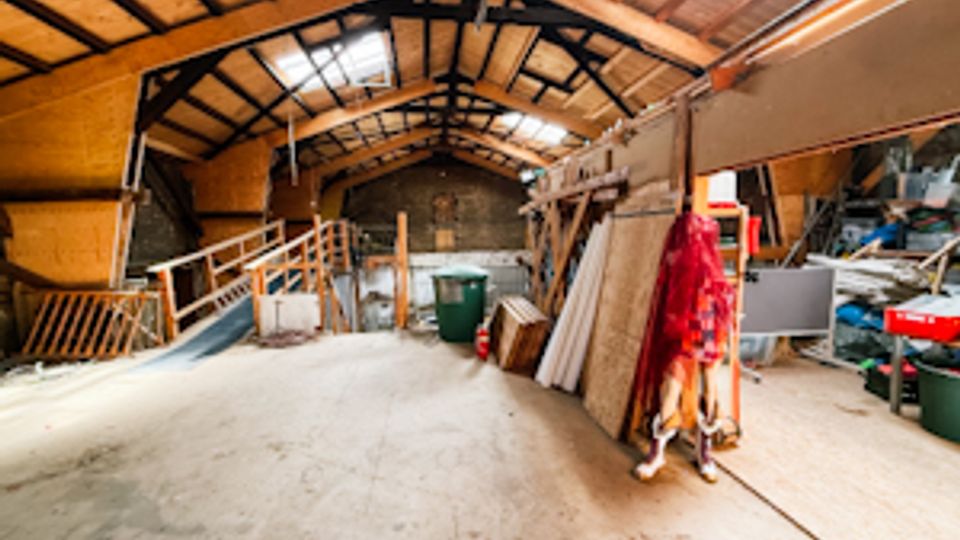 Innenansicht einer großen Scheune oder Lagerhalle mit hohem Satteldach, sichtbaren Holzbalken und Oberlichtern. Auf dem Betonboden stehen Werkzeuge, Regale, Bretter und verschiedene gelagerte Gegenstände entlang der Wände.