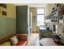 Schmale Küche mit rustikalem Charme, bestehend aus einer grünen Küchenzeile mit Spüle und Regal, einem Holzfußboden im Fischgrätenmuster sowie einem kleinen Essbereich mit rundem Holztisch. Im Vordergrund steht ein orangefarbener Sofa mit bunten Kissen, daneben sind helle Wände und ein großes Fenster sichtbar.